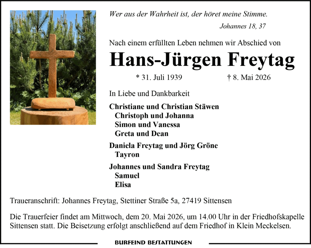 Holz-Kreuz-Grabzeichen auf einem runden Holzteller vor grünem Buschwerk.