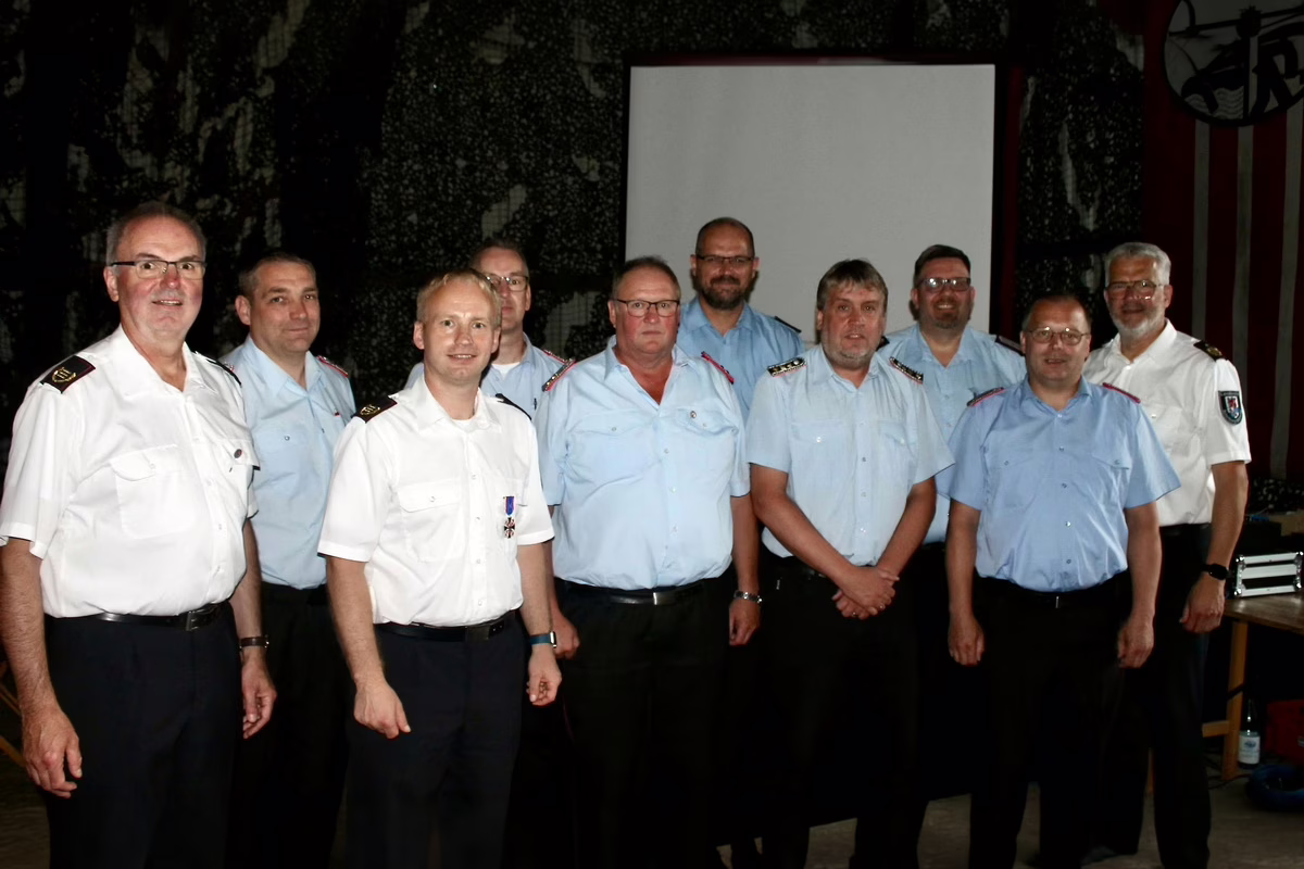 Group of men in light blue and white uniforms posing for a photo indoors, with a projector screen behind them.