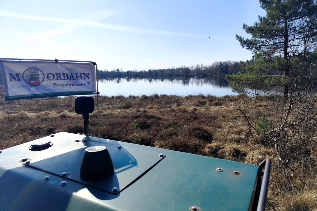 Teal vehicle hood in the foreground with a banner beside a calm lake and pine trees.