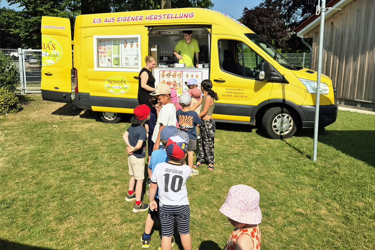 Yellow ice-cream van with a service window, staff serving a line of children outdoors on a sunny day.
