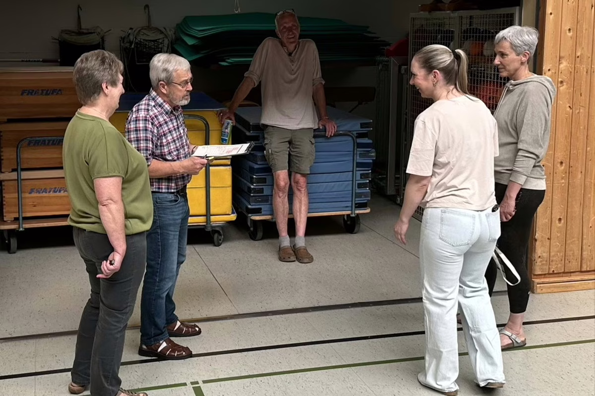 Six adults stand in a gym storage area with stacked mats; one man holds a clipboard and explains something to the group across the room.