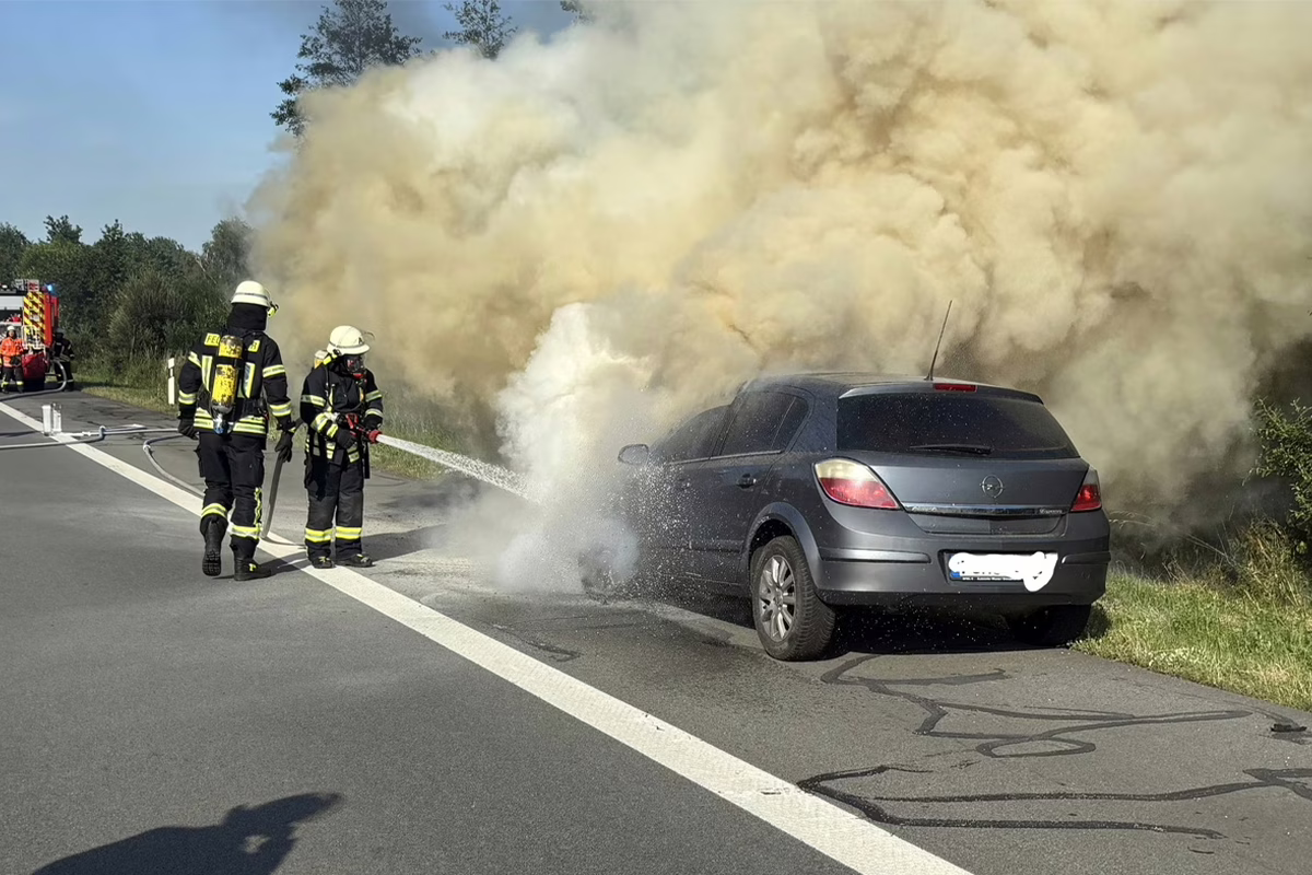 Firefighters hose down a gray hatchback on the roadside as thick yellow smoke pours from the car.
