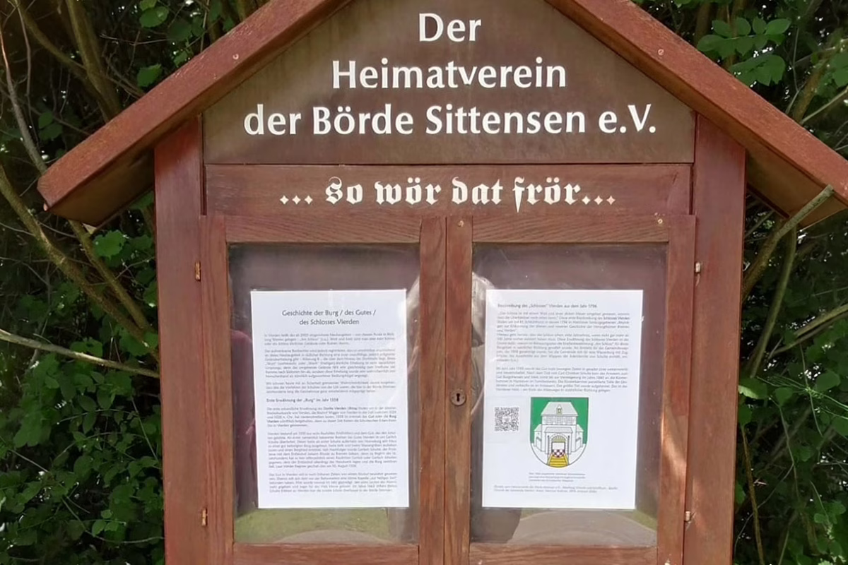 Wooden notice board with a peaked roof displaying the German title 'Der Heimatverein der Börde Sittensen e.V.' and two documents posted behind glass.