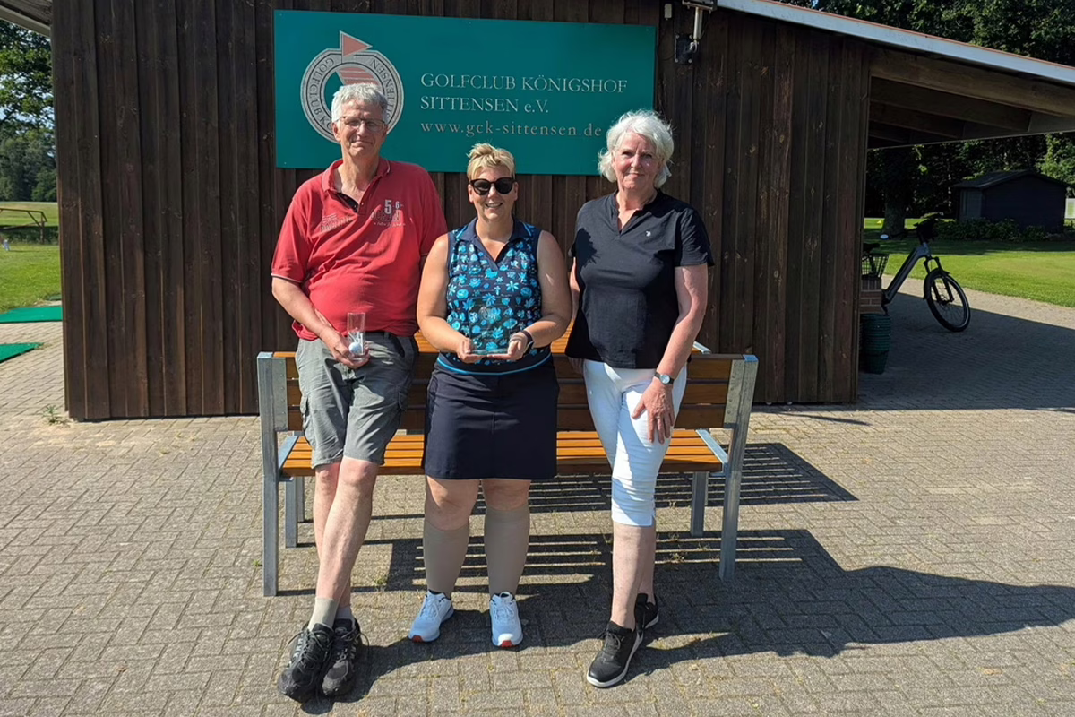 Three adults pose outside a golf club clubhouse; the middle woman holds a small trophy while the others smile.