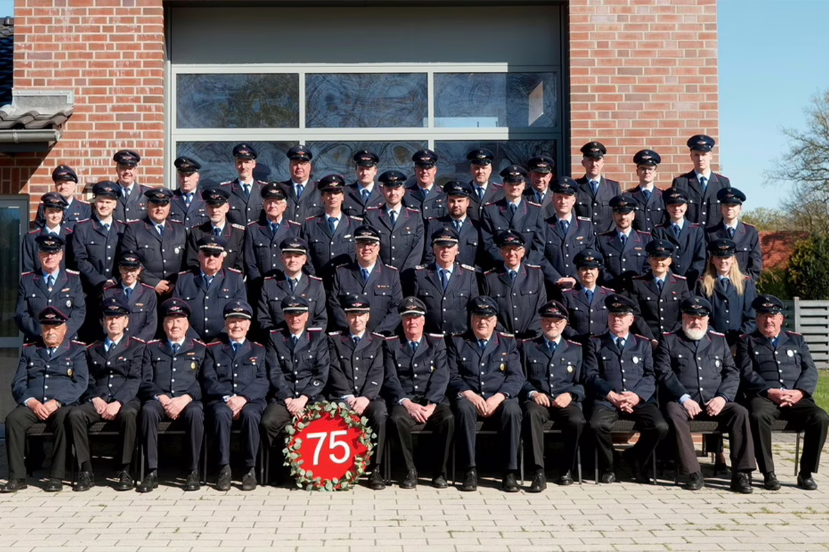 Group of uniformed officers posing for a formal photo in front of a brick building with a red 75 wreath at their feet, commemorating a 75th anniversary or milestone.
