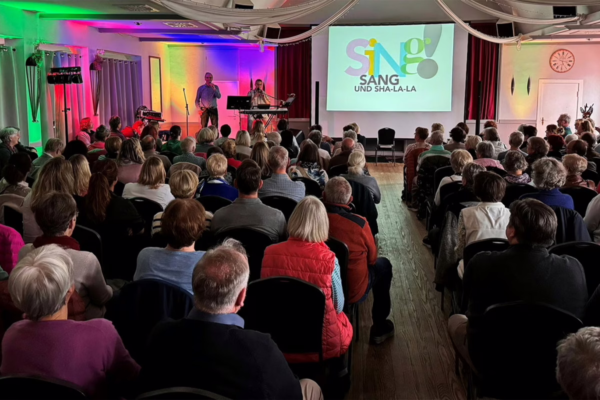 Audience seated in a hall watching a live stage performance; musicians on stage with a keyboard and microphone, projection screen shows a colorful 'SING' logo.