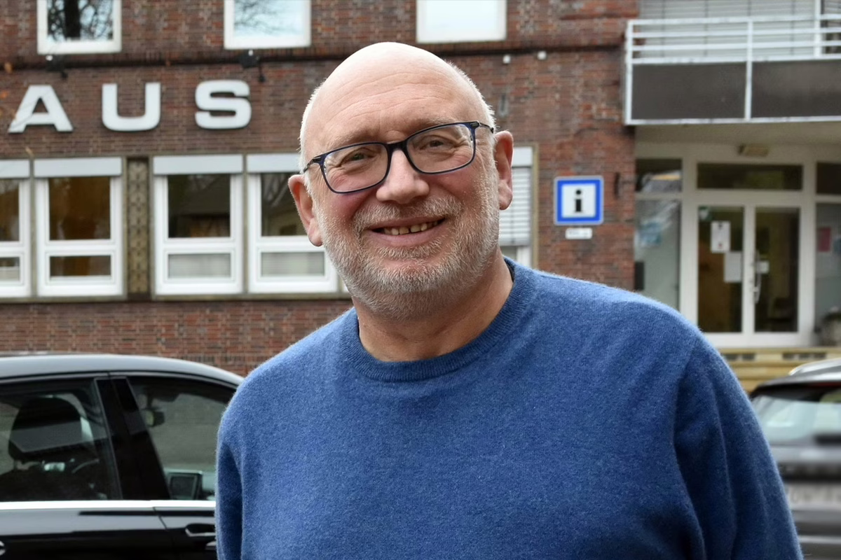 Smiling older man with glasses in a blue sweater outside a brick building with car parked nearby.