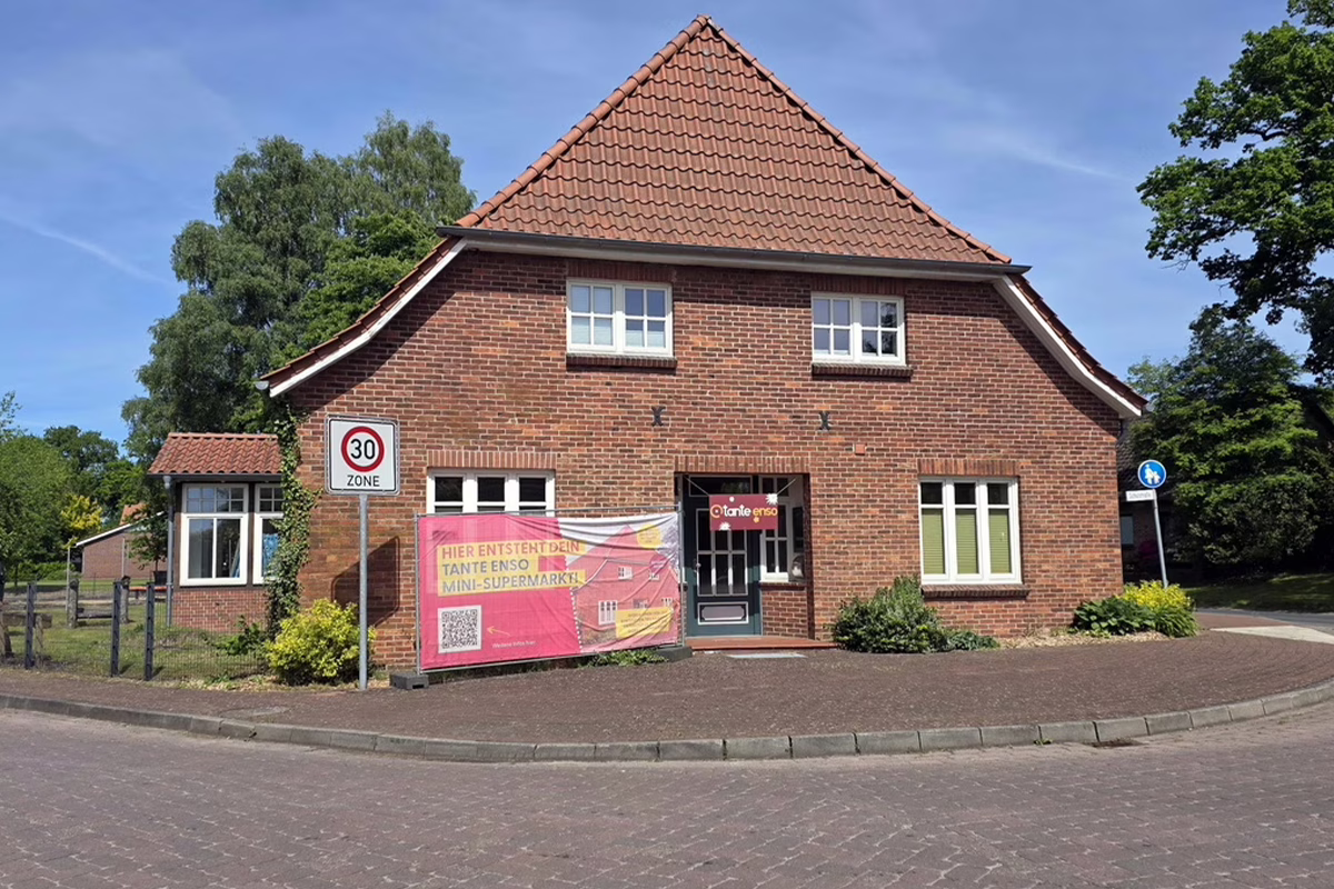 Brick building with a steep red-tiled roof, front entrance, and a pink banner in front advertising a mini-supermarket; 30-zone street nearby.