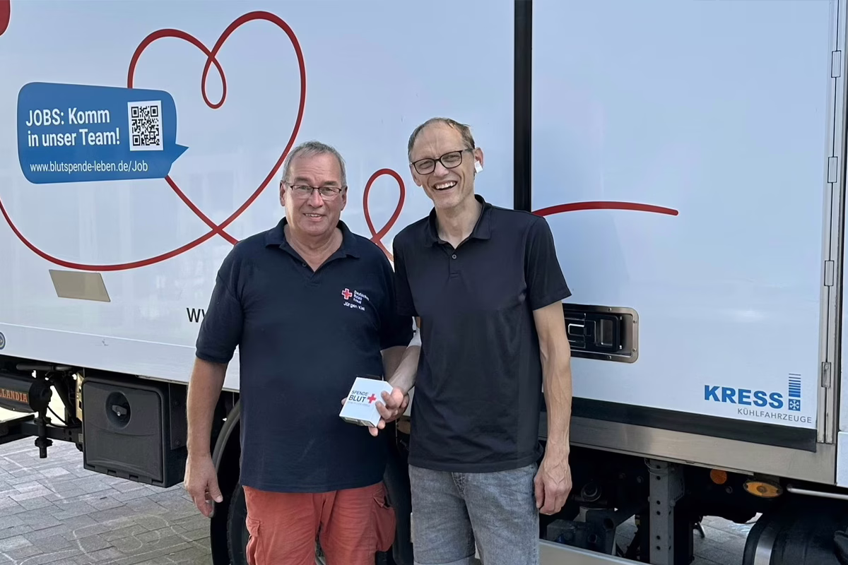 Two men stand side by side in front of a light blue blood donation truck, smiling for the camera; one holds a small donation box labeled Blut. Mom and colleague vibe, casual polo shirts.