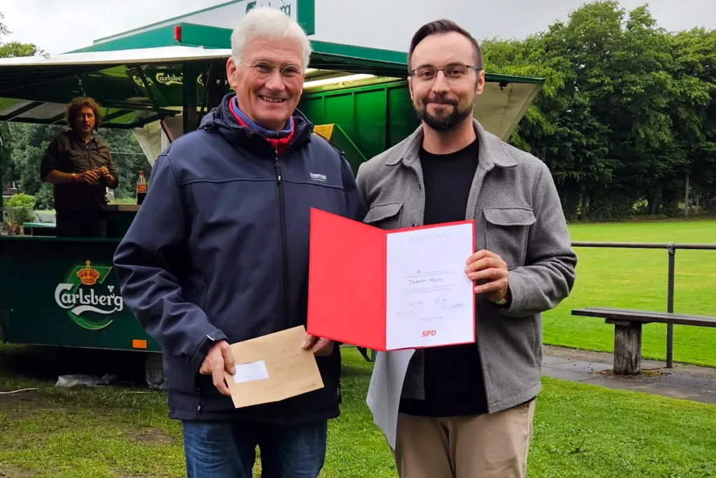 Two men stand in a park; one in a dark jacket holds an envelope while the other presents a red certificate folder with signatures, in front of a green beer trailer.