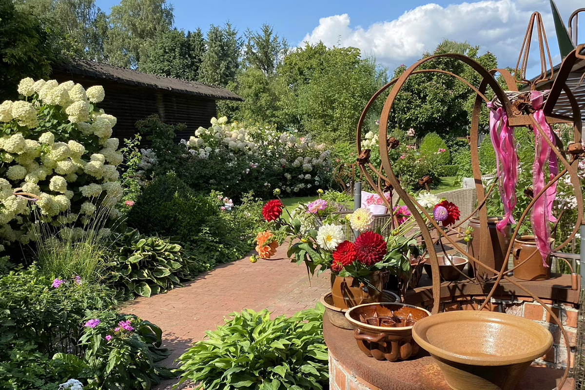 Garden scene with white hydrangeas, a brick path, and a rusted metal sculpture adorned with pink ribbons beside potted flowers.
