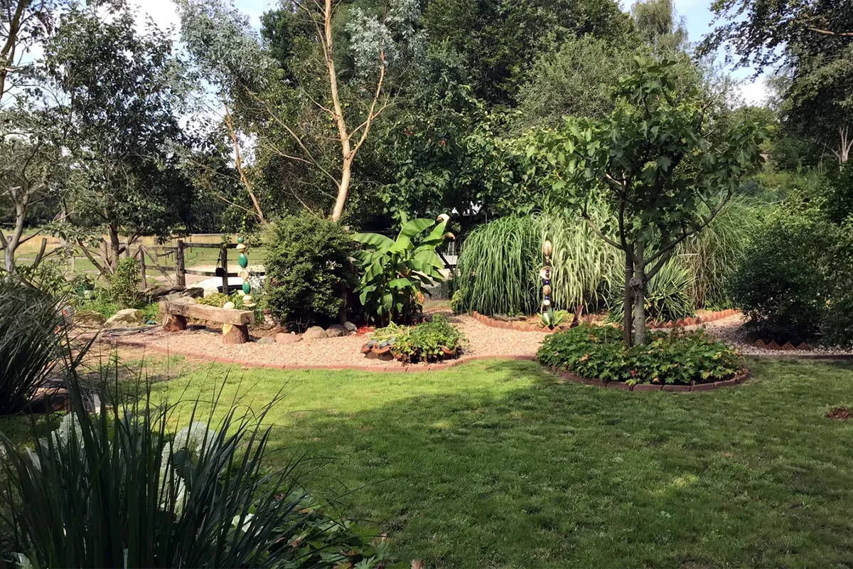 Well-kept garden with a green lawn, trees, a wooden bench, and a circular brick-bordered plant bed beside a colorful stacked-disc sculpture.