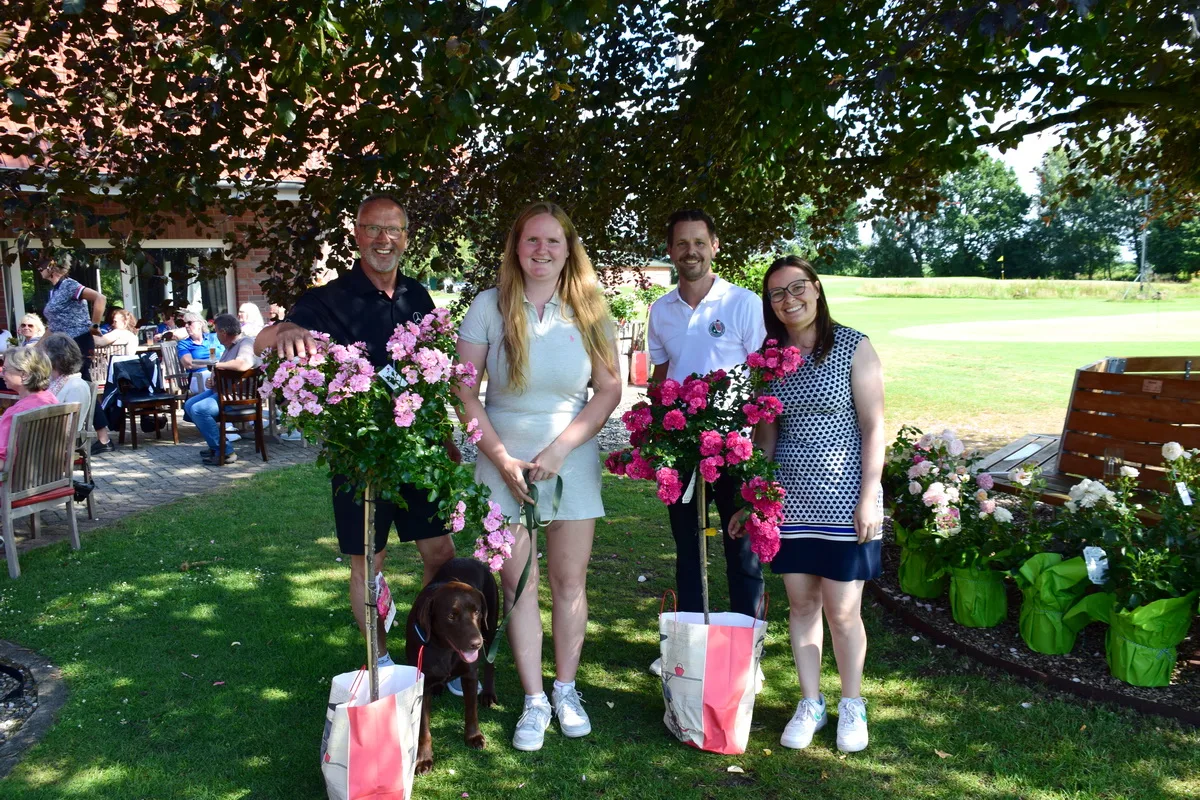 Group of four people posing outdoors under a tree with pink roses on stakes in bags; a brown dog sits by their feet, with guests seated in the background on a patio.