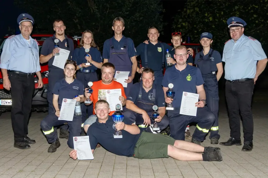 Group of firefighters posing with trophies and certificates after a competition or ceremony, outdoors at night. Front row sits and lies on ground, others stand behind holding awards.