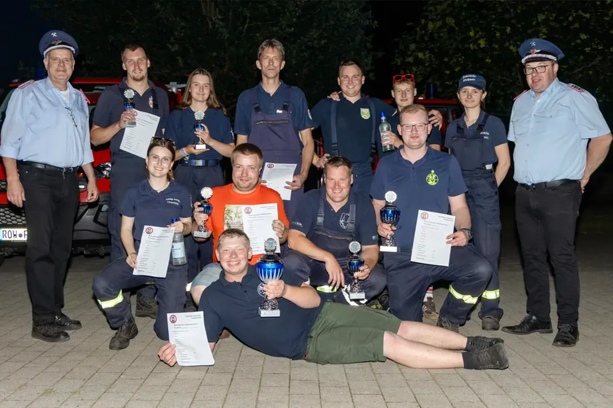 Group of firefighters posing with trophies and certificates after a competition or ceremony, outdoors at night. Front row sits and lies on ground, others stand behind holding awards.
