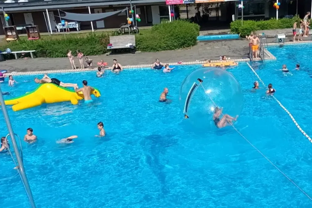 People enjoying a sunny outdoor pool: swimmers around the edge, a large yellow inflatable slide/float on the water, and two people inside a clear inflatable ball.