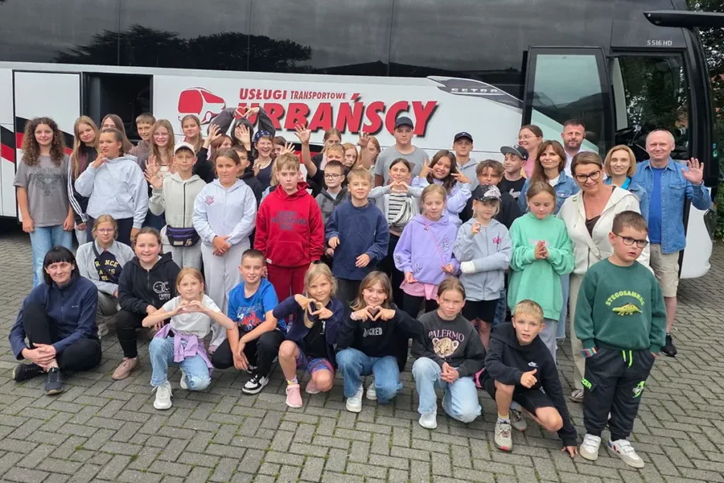 Group of children and a few adults posing for a photo in front of a white touring bus with red lettering, on a paved area, many waving hello.
