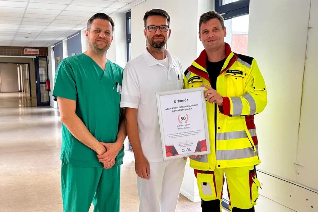 Three healthcare professionals pose in a hospital hallway; green scrubs, white uniform, and high-visibility jacket, holding a framed certificate titled Urkunde (certificate).