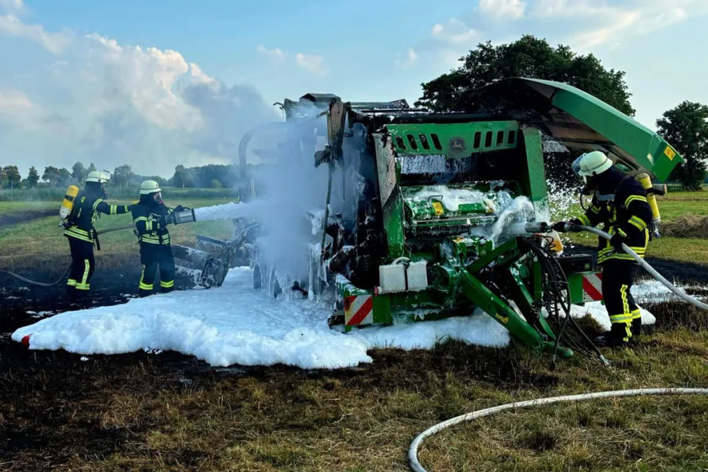 Firefighters in gear spray foam onto a burned green agricultural harvester in an open field.