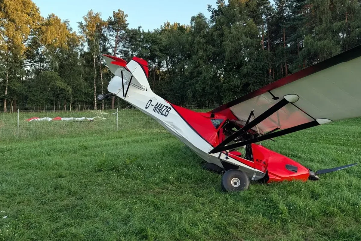Small red-and-white single-engine airplane crashed in a grassy field, with a damaged tail and propeller near trees in the background.