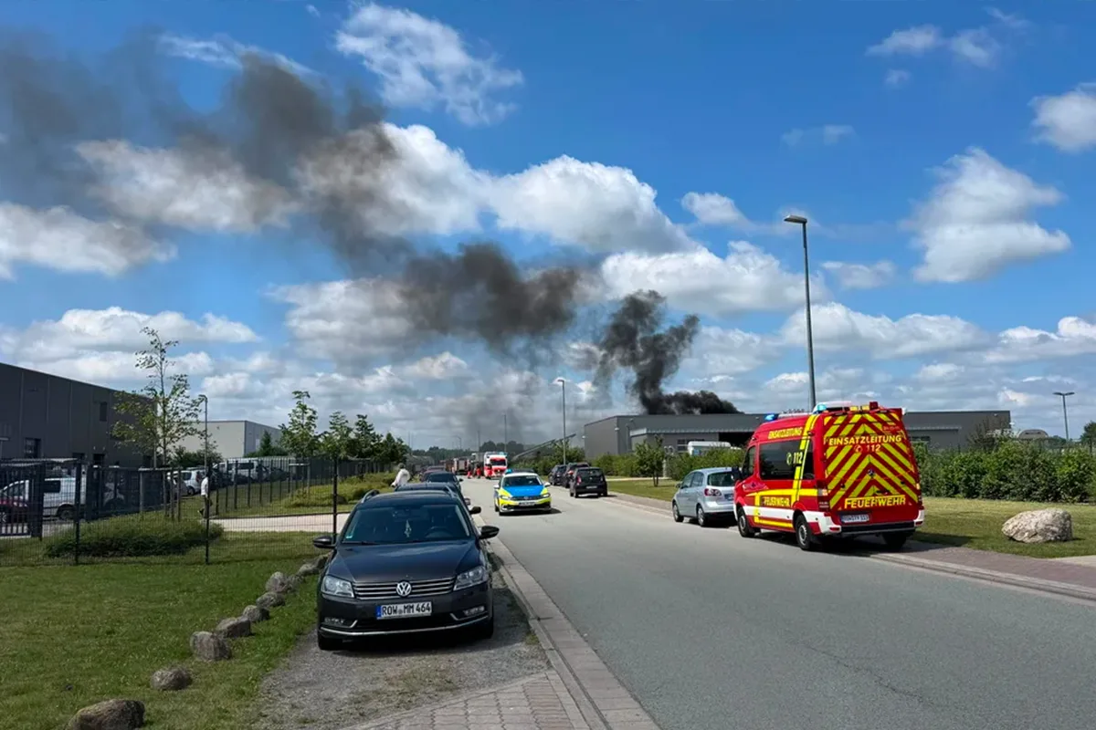 Emergency vehicles respond as dark smoke rises from a building in the distance along a street with parked cars and a blue sky above