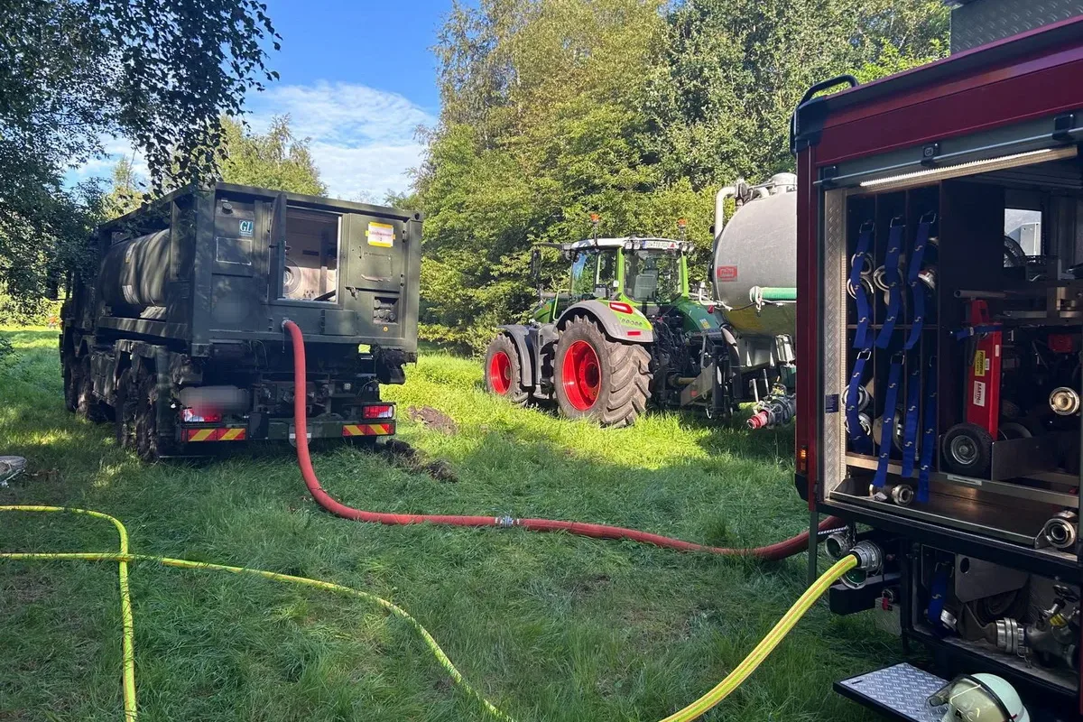 Hose lines connect a tanker trailer to a fire engine in a grassy field on a sunny day.