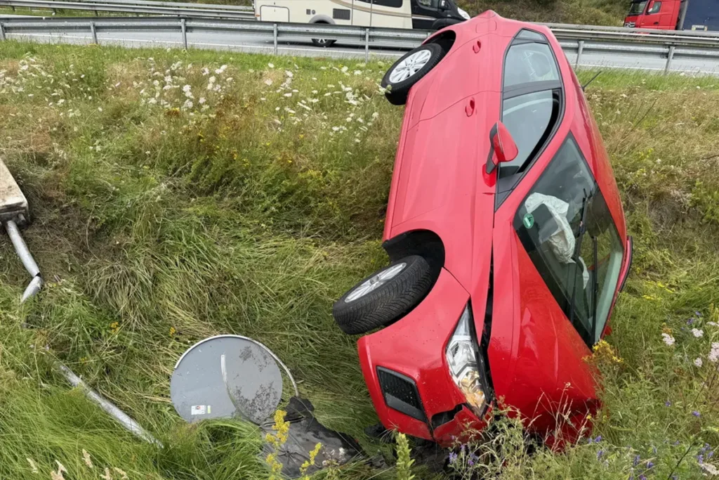 Red hatchback lying on its roof in a grassy ditch beside a highway, with a guardrail in the background and a fallen traffic sign nearby.