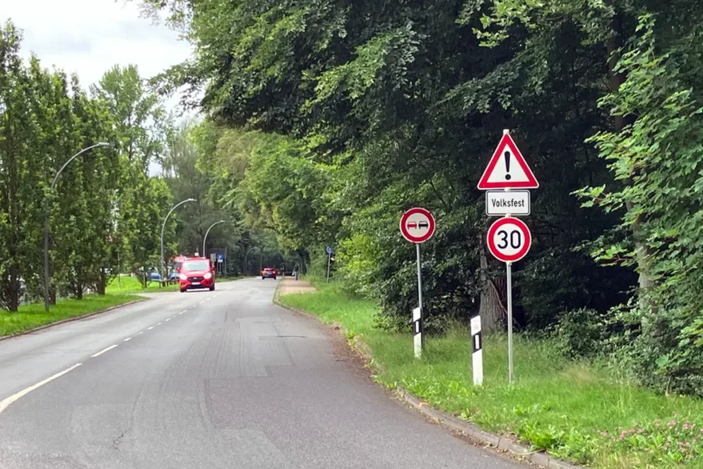 Rural street scene with a red car approaching; road signs on the right warn of a Volksfest and a 30 km/h limit, surrounded by trees and greenery.