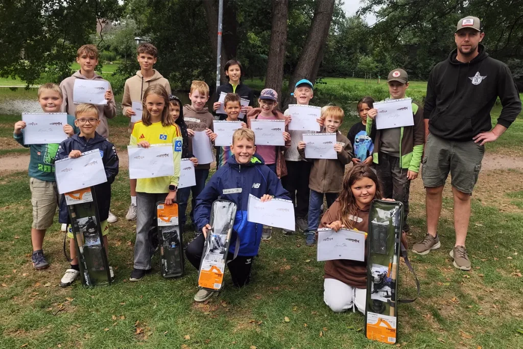 Group of children in a park posing with certificates and prize boxes, with a man in a black hoodie standing to the right.