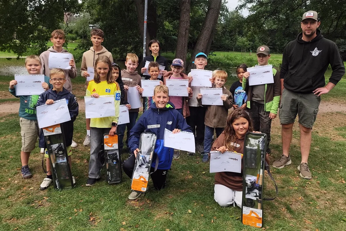 Group of children in a park posing with certificates and prize boxes, with a man in a black hoodie standing to the right.
