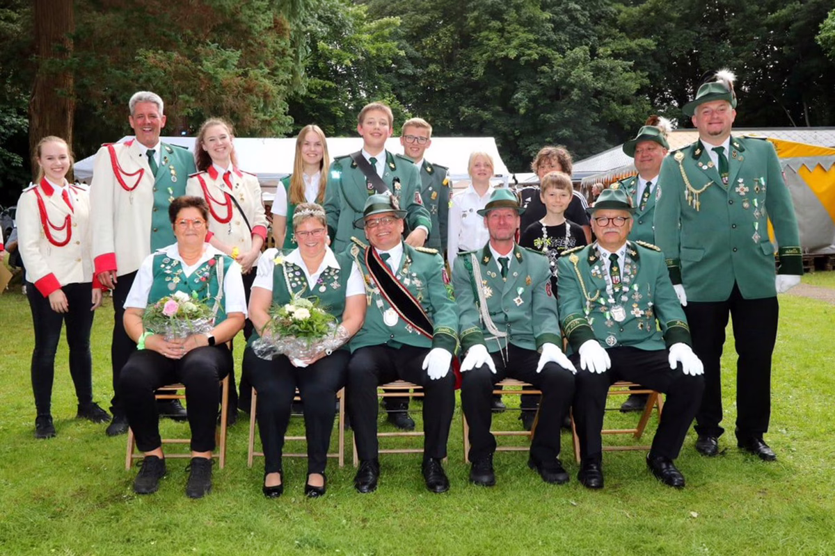 Group of people in green uniforms posing together outdoors, some seated with bouquets in front of a tented event area.