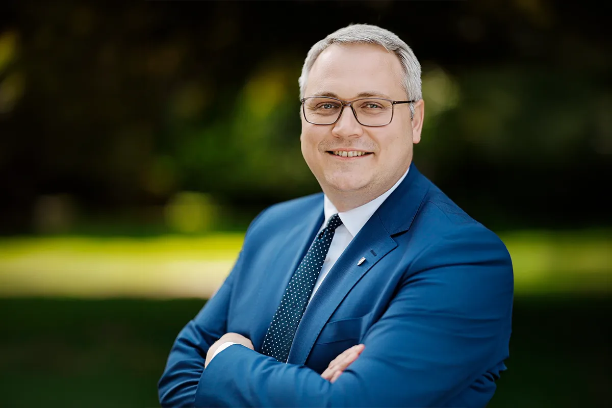 Smiling man in a blue suit and glasses stands outdoors with arms crossed.