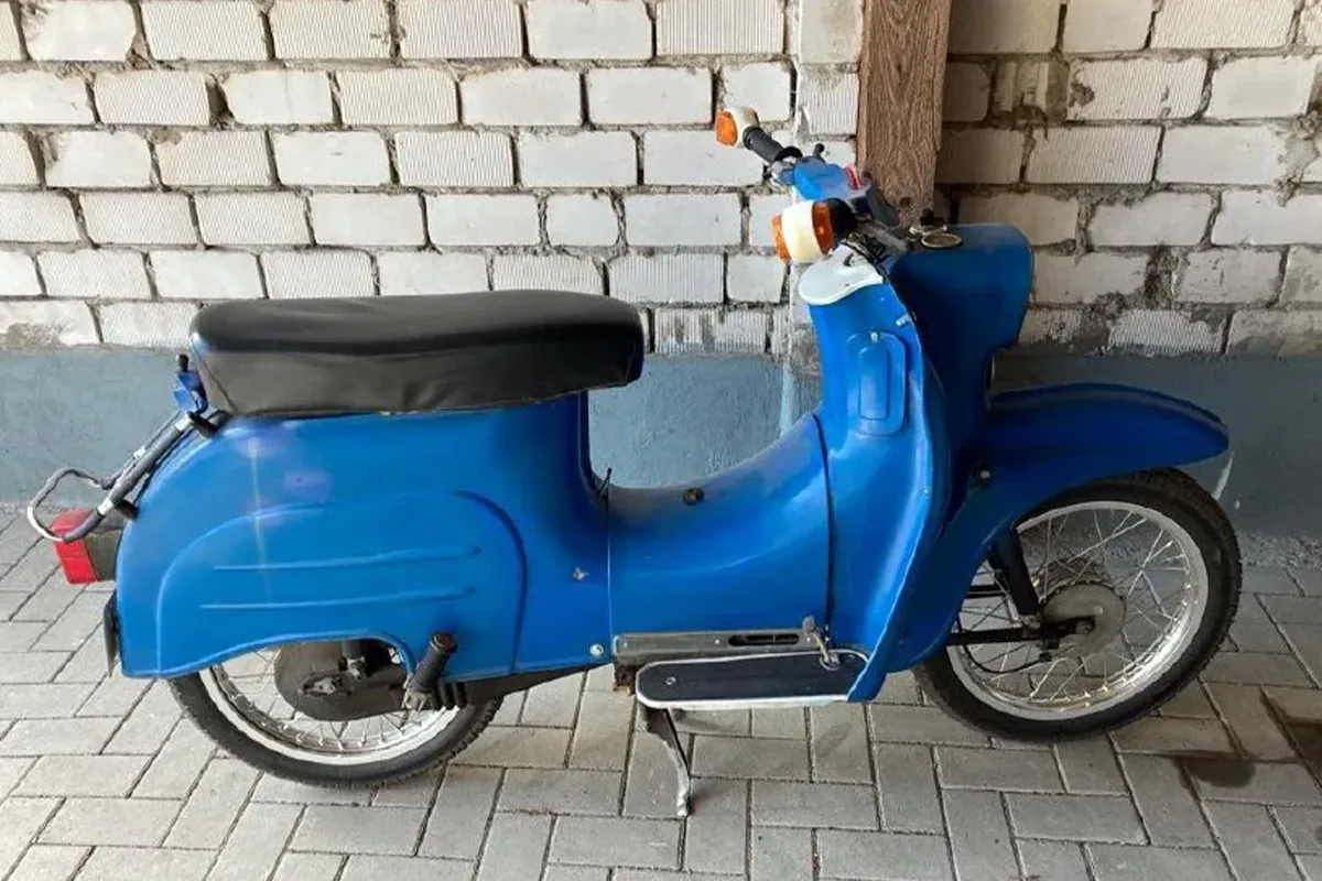 Blue vintage scooter parked indoors against a white brick wall, with a black seat and kickstand.