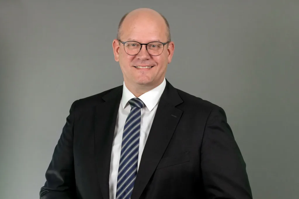 Professional headshot: smiling man in a black suit, white shirt, and striped blue tie against a neutral backdrop.