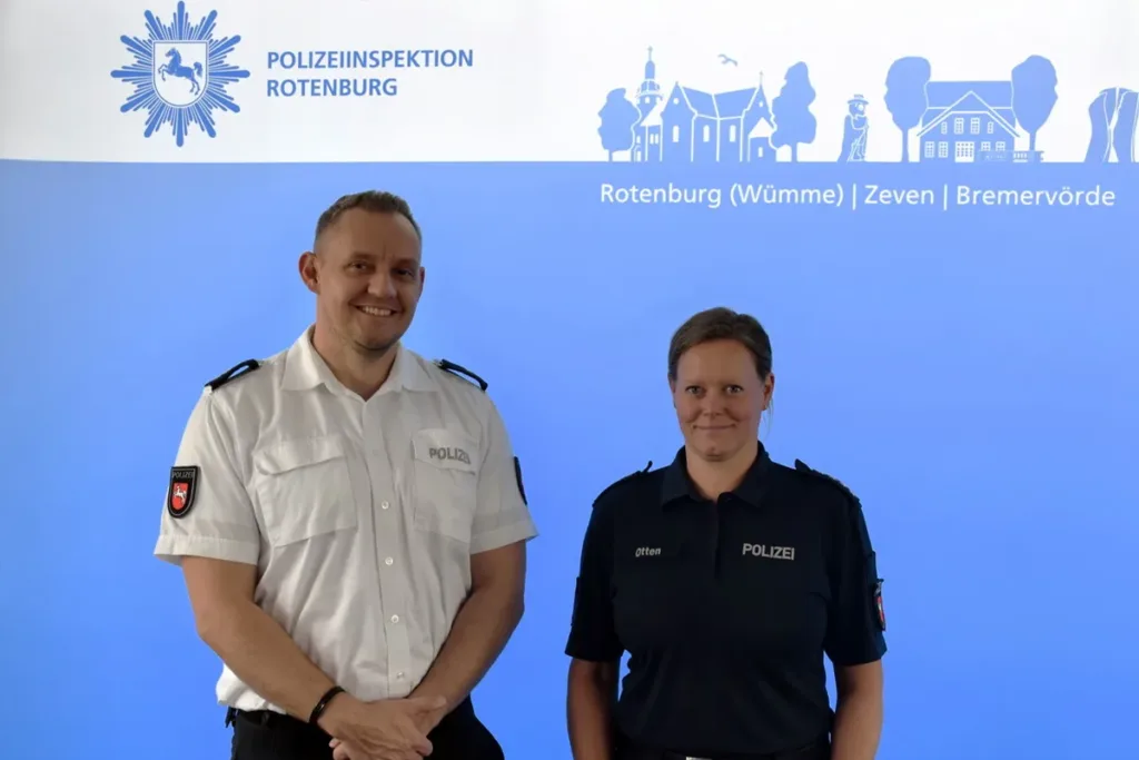 Two police officers stand side by side in uniform in front of a blue backdrop with the Polizeiinspektion Rotenburg logo and crest? (informative)
