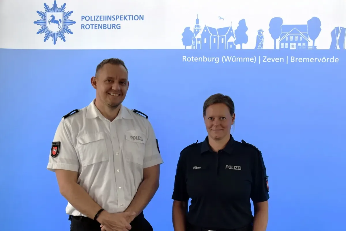 Two police officers stand side by side in uniform in front of a blue backdrop with the Polizeiinspektion Rotenburg logo and crest? (informative)