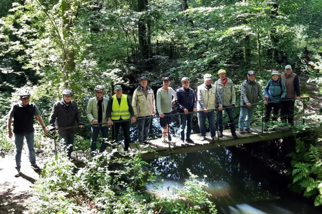 Group of people wearing helmets standing on a wooden bridge over a small forest stream, surrounded by trees.