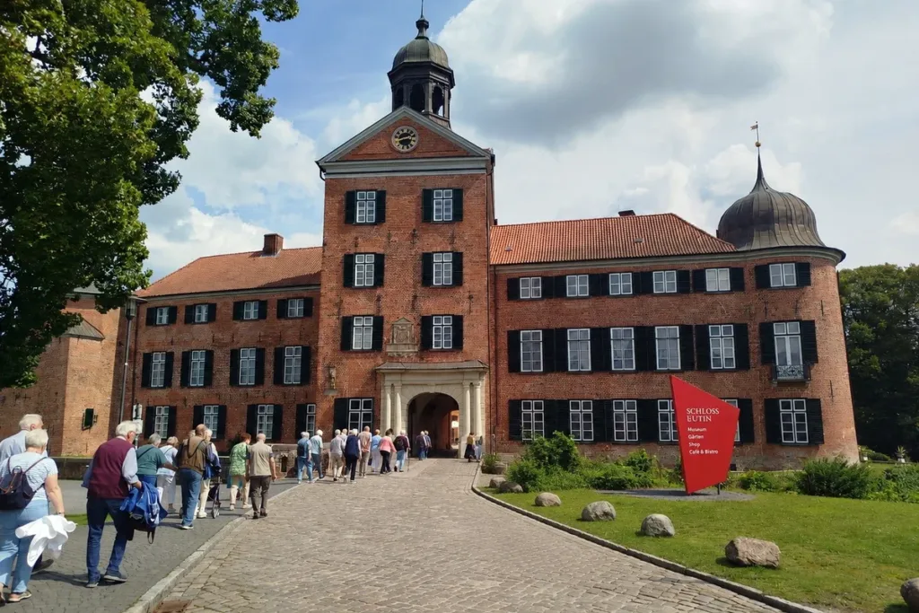 Line of visitors walking along a cobblestone path toward the arched entrance of a red-brick castle with black shutters and a central tower, sunny day.