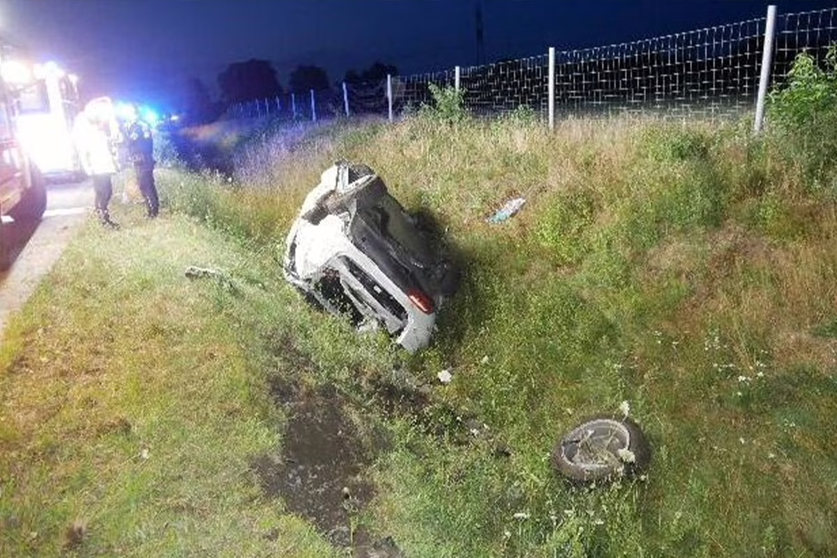 Nighttime crash scene: a white car lies overturned in a grassy ditch beside the road; responders and an ambulance are present.