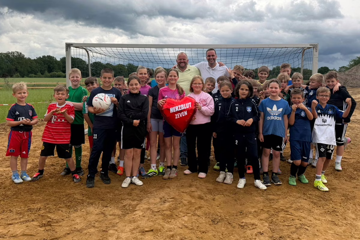 Group of children and two adult coaches pose for a photo on a dirt soccer field in front of a goal; a red heart-shaped balloon reads 'HERZBLUT ZEVEN' held by some children in the center.
