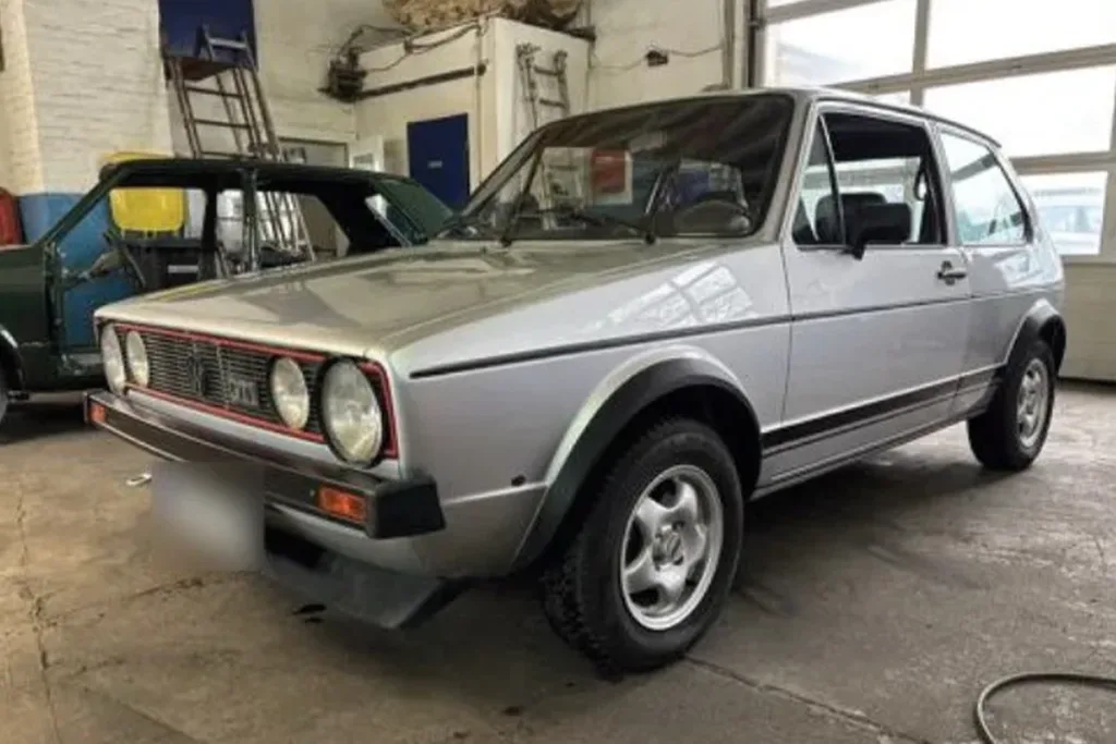 Silver vintage compact car in a workshop, shown from the front‑three‑quarter angle with four round headlights, a red-trimmed grille, and black side moldings; another car and workshop equipment visible in the background.