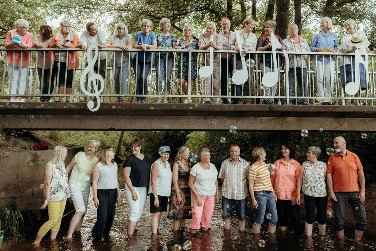 Two rows of adults on a bridge and in a shallow river, smiling on a sunny day, with large musical-note decorations on the railing
