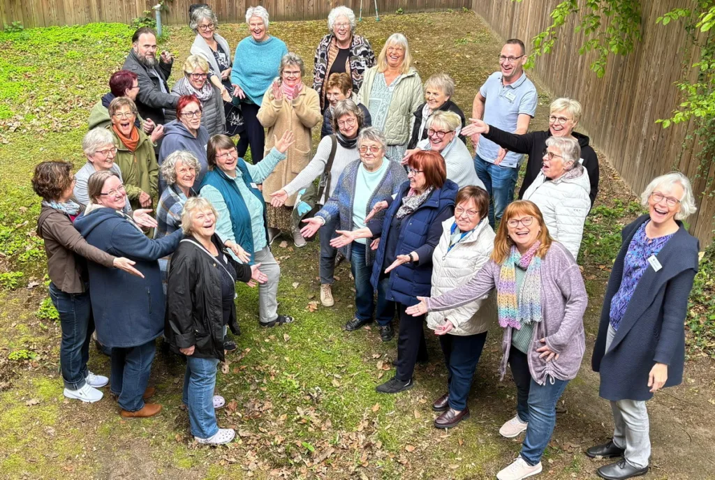 Group of about 25 smiling adults standing in a circle outdoors, arms open, posing for a friendly community photo in a yard with a wooden fence.