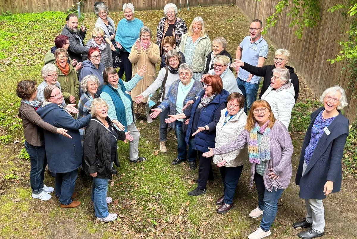Group of about 25 smiling adults standing in a circle outdoors, arms open, posing for a friendly community photo in a yard with a wooden fence.