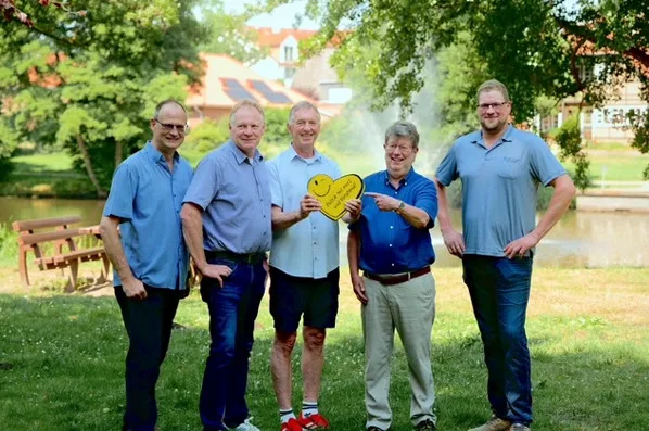 Five men stand on a grassy park near a pond, smiling, with a yellow heart-shaped sign held by the center two men.