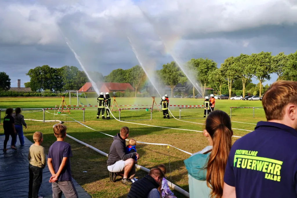 Firefighters in turnout gear spray water across a grassy field while spectators, including children, watch from behind a taped-off area.