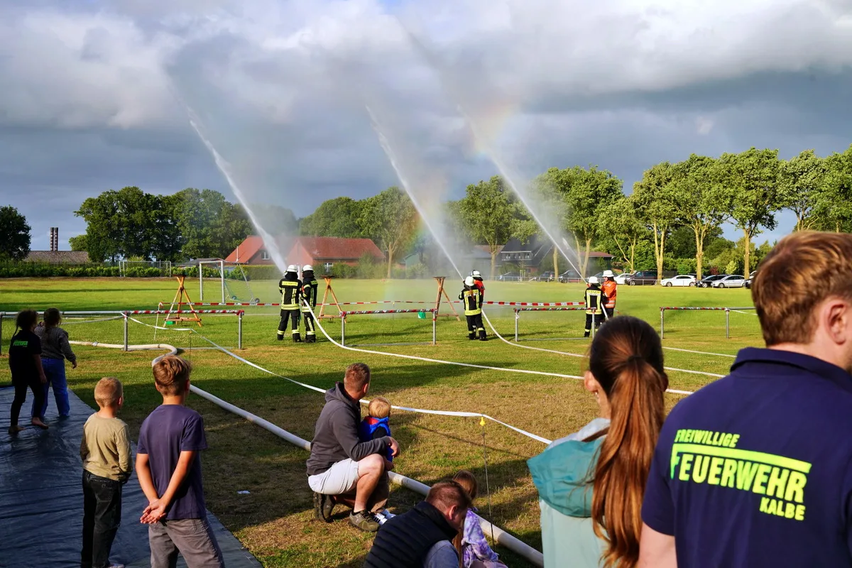 Firefighters in turnout gear spray water across a grassy field while spectators, including children, watch from behind a taped-off area.