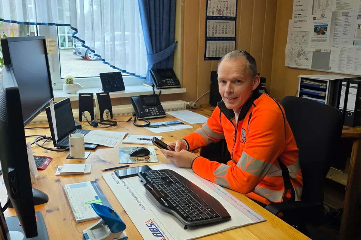 Male worker in a bright orange safety jacket sits at a cluttered office desk with multiple computers, phones, and papers, looking at a smartphone.
