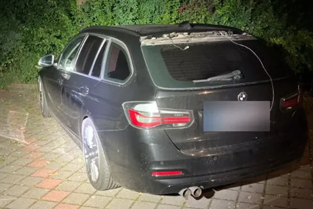 Black BMW SUV parked on a brick driveway at night with a shattered rear window and visible interior damage near the roof area.