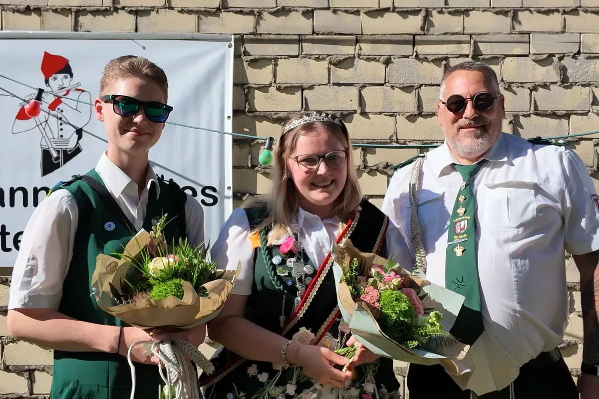 Three people pose outdoors in front of a brick wall, holding bouquets; two youths in traditional dress and an older man in a white shirt and green tie with medals.